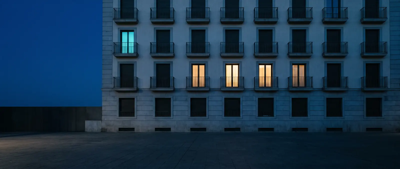 Fachada de un edificio de viviendas al anochecer, con algunas ventanas iluminadas.